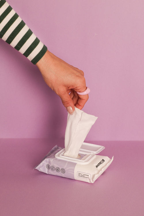 Hand pulling Biolouve V On The Go biodegradable intimate wipe from package against pink background