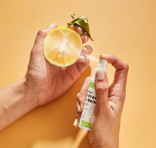 Hands holding Biolouve V Fresh and Ready intimate deodorant mist with halved lemon on orange background