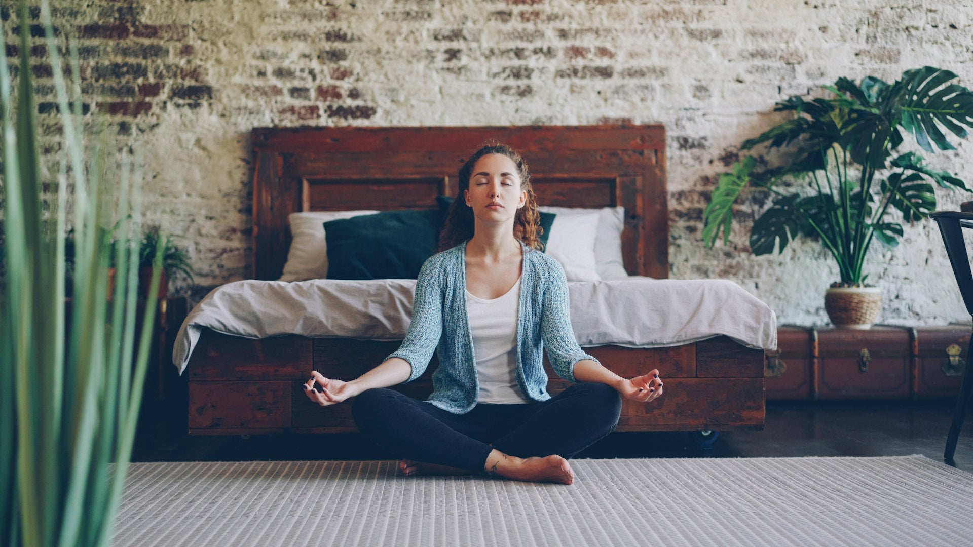 Woman meditating peacefully in bedroom with plants representing daily wellness and self-care routine